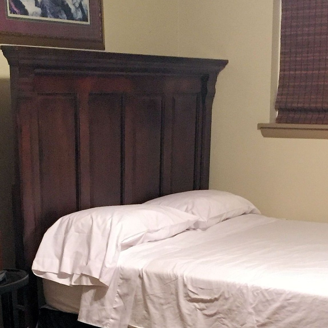 a simple bed, window shade down, small lamp on a side table