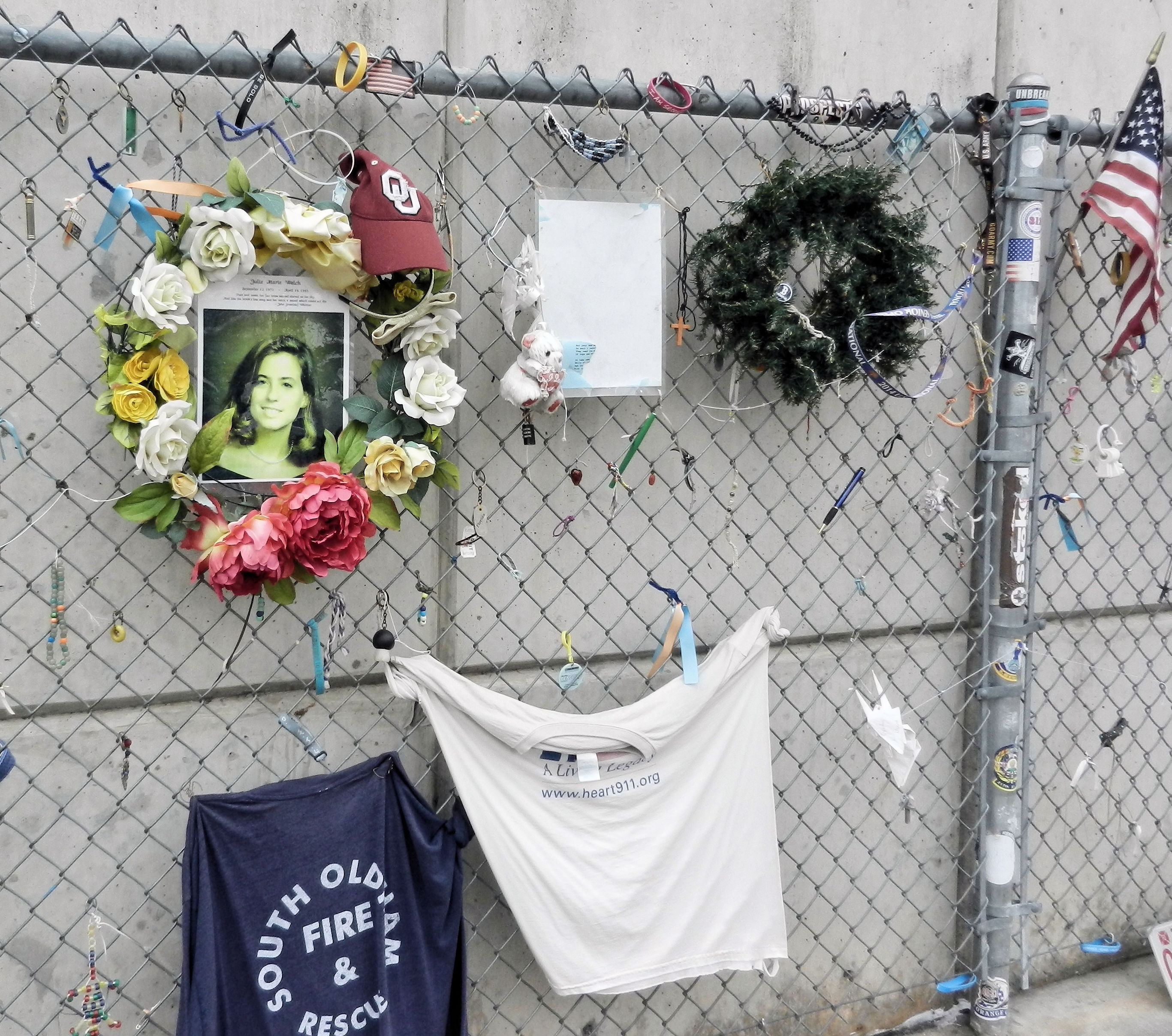 a chainlink fence with mementos-girl photo, teeshirts, wreaths, flag, toy