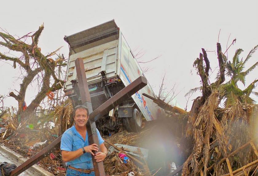 a man with a cross over his shoulder standing in front of a truck and trees damaged in a typhoon