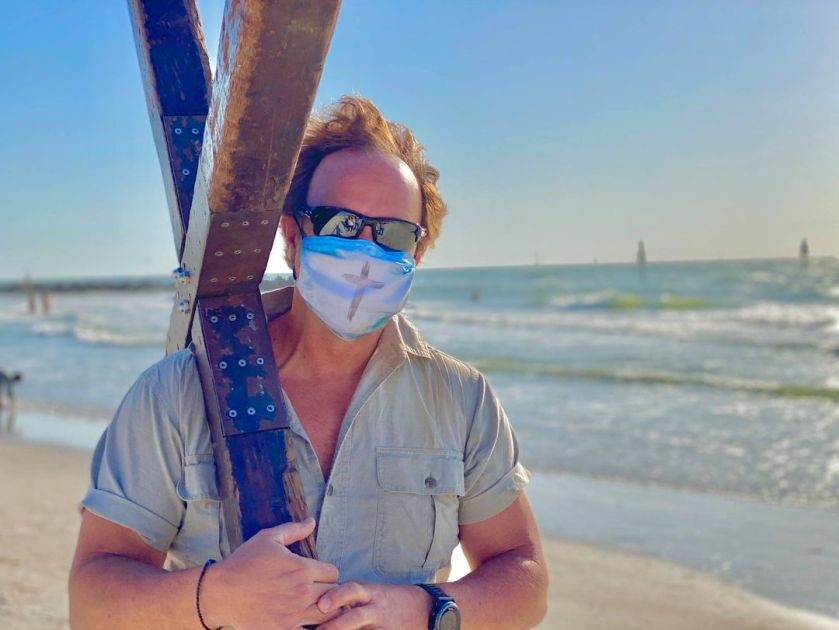 a man on a beach, wearing a mask with a cross drawn on it, and with a cross over his shoulder