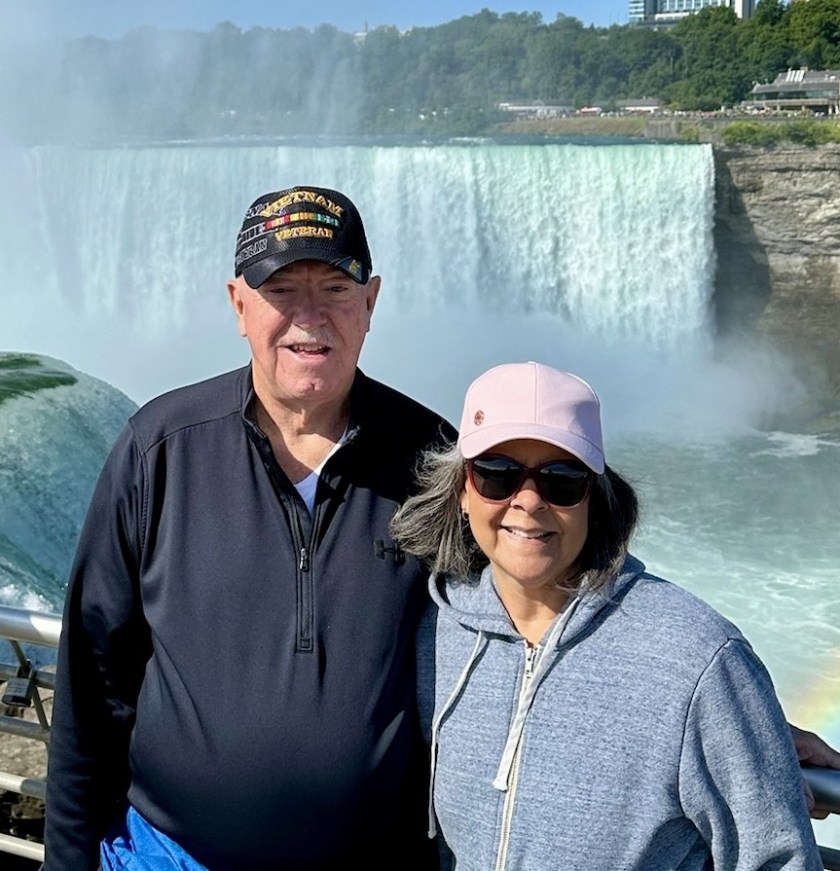 A man and a woman standing together with Niagara Falls in the background, showcasing the beautiful cascading water and mist.